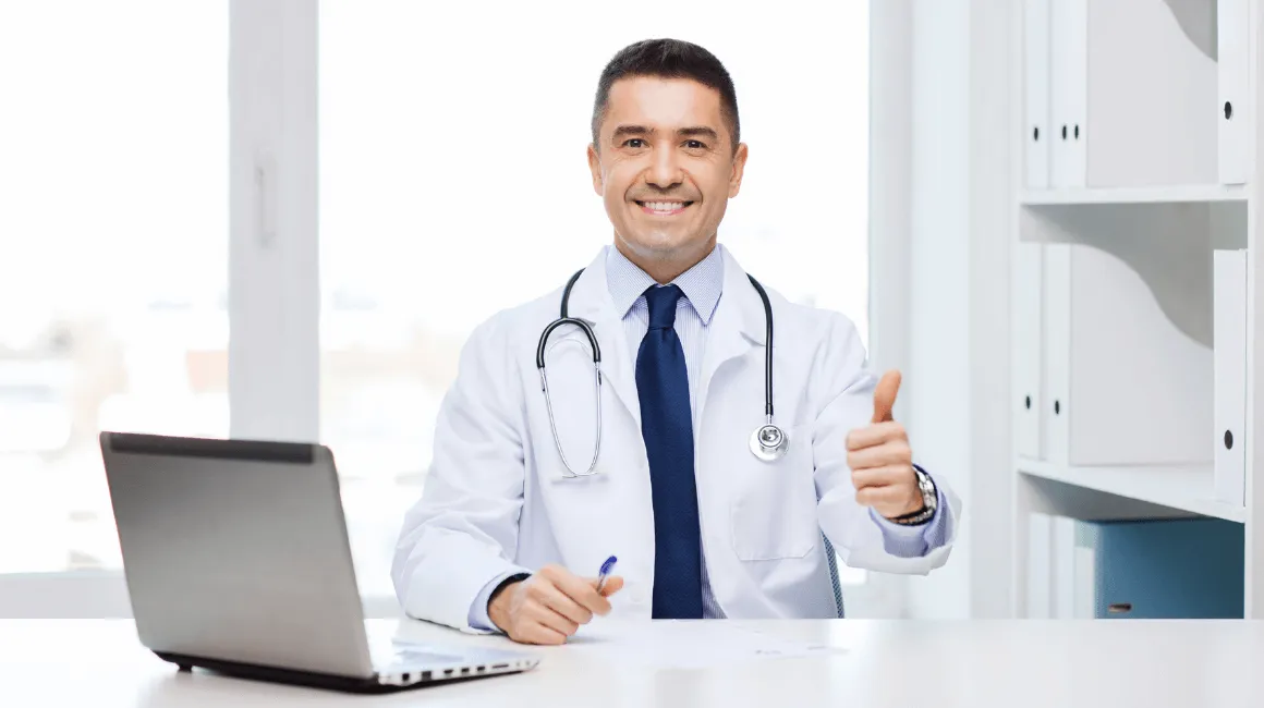 Lekarz w białym fartuchu z stetoskopem na szyi siedzi przy biurku z laptopem i pokazuje gest „OK” kciukiem, uśmiechając się do kamery